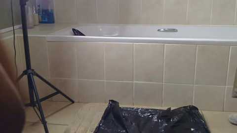 Media: Video of a beige-tiled bathroom with a white bathtub, a black trash bag, and a tripod standing against the wall.