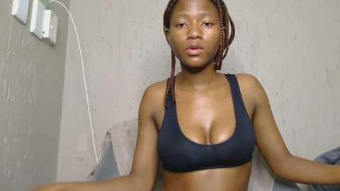 Media: Video of a young Black woman with braided hair in a black sports bra, sitting against a plain, light-colored wall with a white outlet and cord on the left.