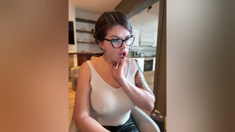 Media: Video of a woman with short dark hair, wearing black-framed glasses, a white ribbed tank top, and black pants, seated in a modern kitchen with light wood cabinetry, touching her neck, looking surprised.