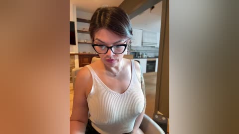 Media: Video of a fair-skinned woman with dark hair, wearing black-rimmed glasses, a white ribbed tank top, and black shorts, sitting in a modern kitchen.