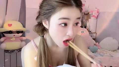 A video of an Asian woman with light skin and long brown hair in pigtails, eating ramen in a pastel-colored room with plush toys, a Hello Kitty doll, and a yellow bear.