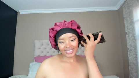 Media: Video of a smiling, light-skinned woman with medium-length hair wearing a pink satin bonnet, using a smartphone while topless in a modern bedroom.