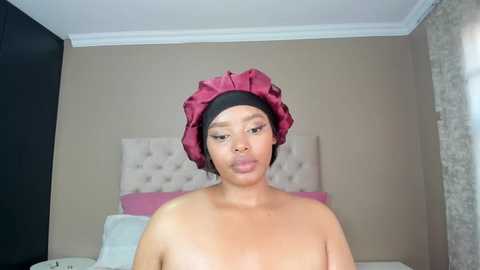 Media: A video of a topless woman with a medium brown complexion and short black hair, wearing a large, ruffled, maroon turban, sitting in a bedroom with beige walls, a tufted headboard, and pink bedding.