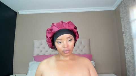 Media: A video of a topless woman with light brown skin, wearing a large red turban and a black headscarf, sitting on a bed with a beige tufted headboard.