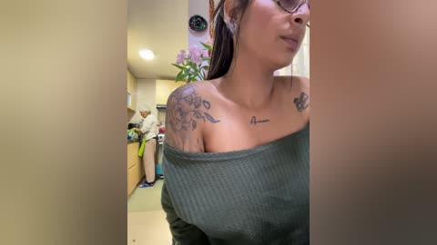 Media: Video of a tattooed woman with glasses, wearing a green off-the-shoulder top, standing in a modern, brightly lit room with a potted plant in the background.