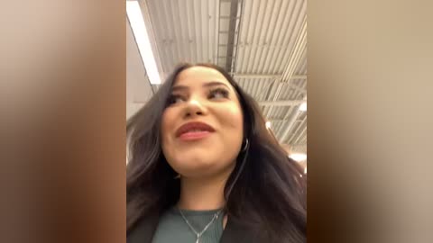 Media: A video of a smiling Latina woman with long, wavy black hair, wearing a green top and a silver necklace, standing in an airport terminal with a corrugated ceiling and fluorescent lights.