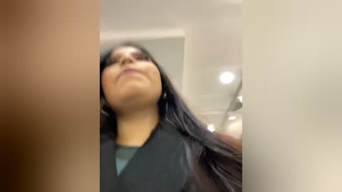 Media: A blurry video shows a woman with long black hair, wearing a dark jacket and green shirt, looking up from below, in an indoor setting with white walls and ceiling lights.