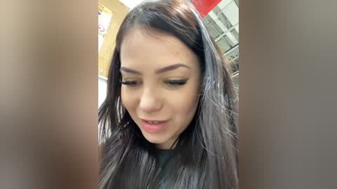 Media: Video of a young woman with long, straight black hair and light skin, wearing a black top, looking down. Background includes a wall with posters and a window.