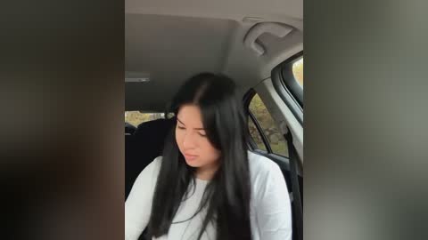 Media: Video of a young East Asian woman with long black hair, wearing a white long-sleeved shirt, seated in the backseat of a car, looking down with a neutral expression.