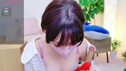 Media: Video of an East Asian woman with short brown hair, wearing a light blue mask and a white shirt, kneeling on a beige couch. She holds a red object near her chest. Background features a blue cushion, potted plant, and beige walls.