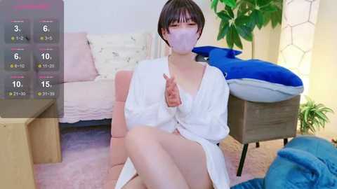 Media: A video of a young woman with short black hair, wearing a white robe, a mask, and gloves, sitting on a pink couch in a room with a blue blanket and a green plant.