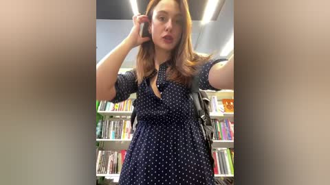 Media: A video of a young Caucasian woman with light skin and shoulder-length brown hair, wearing a navy blue polka-dotted blouse and black backpack, standing in a brightly lit library, holding a phone to her ear.