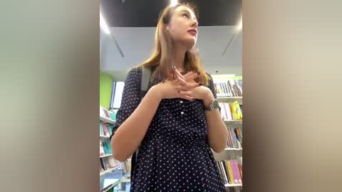 Media: Video of a young woman with light skin, straight brown hair, wearing a navy blue dress with white polka dots, standing in a brightly lit library, gazing upward, holding her chest, surrounded by bookshelves.