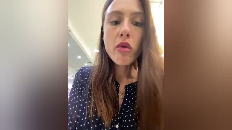 Media: Video of a Caucasian woman with long brown hair, wearing a black blouse with white polka dots, making a kissy face. The background shows a modern, bright kitchen with white walls and recessed lighting.