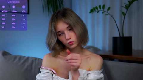 Media: Video of a young woman with short, light brown hair, wearing a white off-shoulder top, making a pouty face. Background features a modern living room with a potted plant and a digital clock.