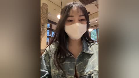 Media: Video of an East Asian woman with long, straight black hair, wearing a white mask and a denim jacket, standing indoors against a wall with decorative, beige, patterned mural.