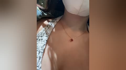 Media: Video of a woman's neck and chest, wearing a white bra with a small, red pendant necklace. Her skin is light, and she is lying on a patterned, white and black fabric.