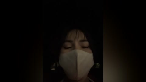 Media: Video of a woman with closed eyes, wearing a white surgical mask and dark hair, set against a dim, dark background.