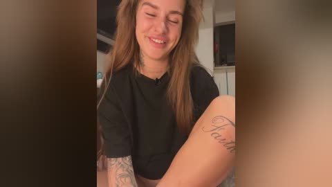 Media: Video of a smiling young woman with long brown hair, wearing a black shirt, revealing a tattoo on her arm. She's indoors, with blurred background.