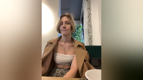 Media: Video of a blonde woman with short hair, wearing a beige trench coat and a white crop top, sitting at a table in a dimly lit caf\u00e9.