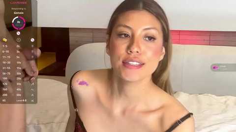 Media: Video of a light-skinned woman with straight, light brown hair, wearing a red and black off-shoulder top, sitting on a bed. Background includes a wooden headboard, a digital clock, and a pink device.