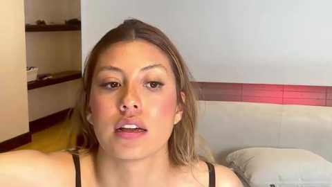 Media: Video of a young woman with light skin, light brown hair, and brown eyes, wearing a black tank top, sitting on a white couch in a modern, minimalistic room with beige walls and a red accent strip.