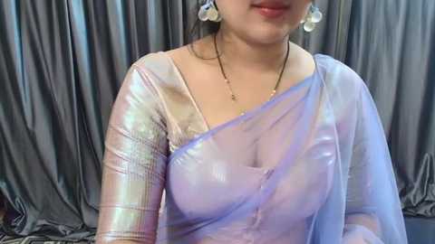 Media: A video of a South Asian woman with medium skin tone, wearing a sheer, pastel blue and silver sequined saree with a plunging neckline, revealing a hint of her ample cleavage. She has dangling earrings and a delicate necklace. Background features draping gray satin fabric.