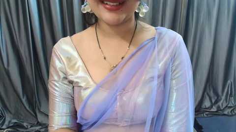 Media: Video of a smiling woman with fair skin, wearing a sheer, pastel lavender sari with silver threads, over a white blouse, against a grey satin backdrop. She has white pearl earrings and a gold necklace.