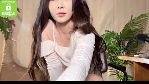 Media: Video of an Asian woman with long black hair, fair skin, and slender physique, wearing a white off-shoulder knit top. Background includes green plants, wooden desk, and beige curtains.