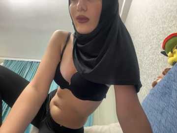 Media: Video of a light-skinned woman with a slender physique, wearing a black hijab and black lingerie, sitting in a room with teal curtains and a stuffed toy.