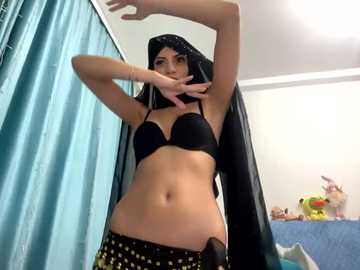 Media: Video of a woman with long black hair, wearing a black bra and patterned skirt, striking a pose in a bedroom with teal curtains, blue bed, stuffed toys, and a white ceiling.