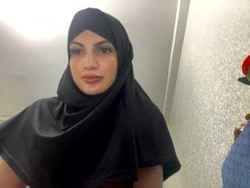 Media: A video of a woman wearing a black hijab and a red dress, standing in a bathroom with a beige textured wall and a red towel hanging on the right.