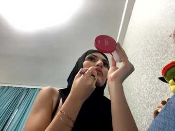 Media: Video of a woman with a fair complexion, wearing a black hijab, applying makeup with a red compact mirror, in a room with a textured white wall, teal curtains, and a colorful toy.