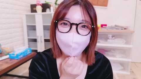 Media: Video of an Asian woman with straight, shoulder-length brown hair, wearing glasses, a white mask, and a black top, standing in a modern, white-walled room with shelves and a table.