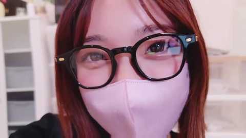 Media: Video of a young woman with long, auburn hair, wearing black-framed glasses and a white surgical mask, standing indoors with shelves and a white door in the background.