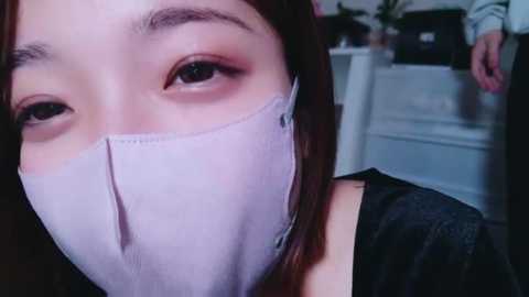 Media: A close-up video of a young Asian woman with fair skin, wearing a light blue surgical mask and dark eyeliner, in a blurred, indoor setting.