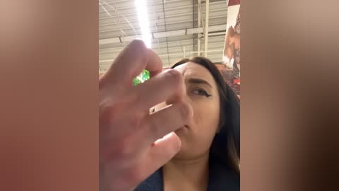 Media: Video of a young woman with long dark hair, wearing a dark top, examining a green gemstone ring on her finger. Background shows a warehouse with industrial ceiling lights and other partially visible people.