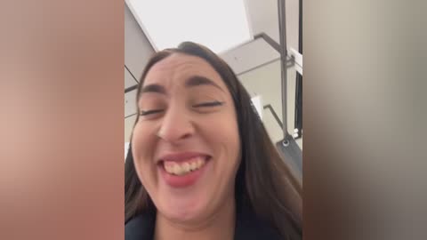 Media: Video of a young woman with long, dark hair, smiling broadly with eyes closed, in a bathroom with beige walls and metal fixtures, including a toilet.