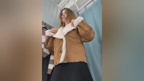 Media: Video of a woman in a beige fur-trimmed jacket and black skirt, standing in a brightly lit room with blue curtains and a striped blanket on a white metal rack.