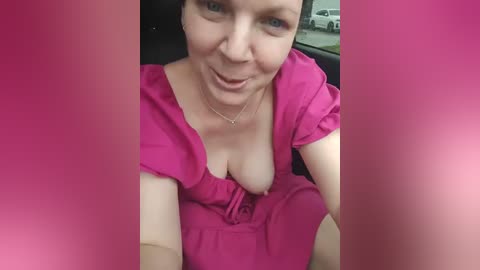 Media: Video of a fair-skinned woman with blue eyes, wearing a low-cut pink dress, revealing cleavage, sitting in a car. Background includes blurred outdoor scenes.