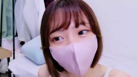 Media: Video of an Asian woman with straight brown hair, wearing a white surgical mask, standing in a hospital room with white walls and a bed.