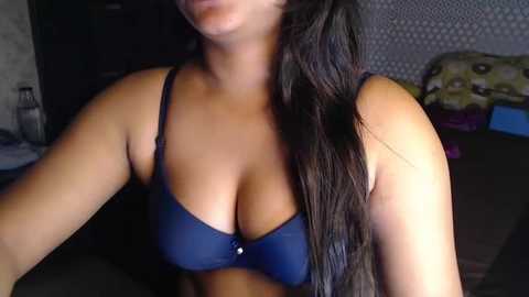 Media: Video of a woman with long, dark hair, wearing a dark blue bra with small, round cups, seated on a bed with a patterned pillow. The background features a dark, possibly wooden, wardrobe and a textured wall.