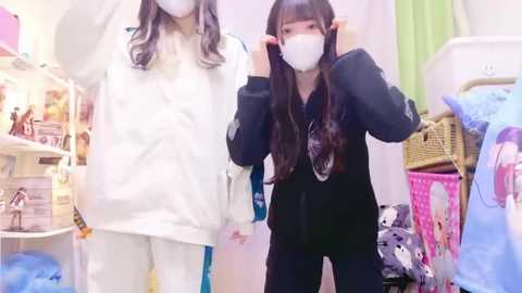 Media: Video of two women in white hazmat suits and face masks, one adjusting her hair, in a cluttered, brightly lit room with toys and laundry.