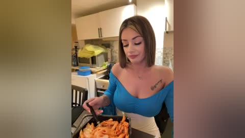 Media: Video of a light-skinned woman with shoulder-length brown hair, wearing a blue off-shoulder top, seated at a kitchen table, eating pasta from a black pan. The background features a modern kitchen with white cabinets and stainless steel appliances.