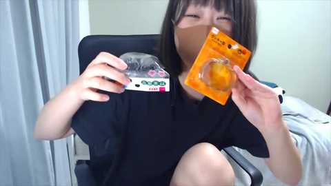 Media: Video of a young Asian woman with straight brown hair, wearing a black shirt, sitting in a black chair, holding a condom packet and a small box.