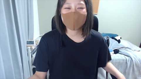 Media: Video of an Asian woman with shoulder-length black hair, wearing a black T-shirt and brown medical mask, seated in a hospital room with a bed, gray curtains, and a wooden door in the background.