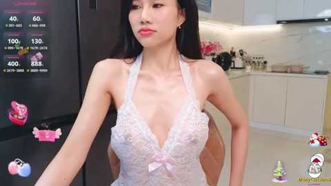 Media: Video of a slim Asian woman in a white lace bralette, standing in a modern kitchen. Background shows a fridge, counter, and white cabinets.