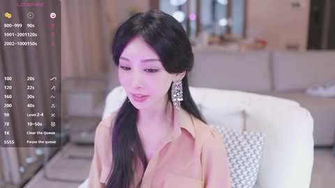 Media: A video of a young East Asian woman with long black hair, wearing a beige blouse and large earrings, sitting on a white couch.