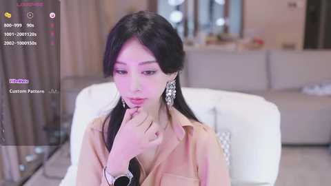 Media: Video of a young Asian woman with long black hair, wearing a beige blouse, seated on a white chair, indoors, with soft lighting and blurred background.