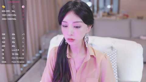 Media: Video of a young East Asian woman with long black hair, wearing a beige blouse, sitting on a white couch in a modern living room.